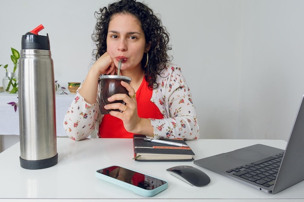 Foto jovem mulher trabalhadora latina em curlers sentada no escritório com telefone portátil e caderno na mesa ela está bebendo mate durante sua pausa de trabalho imagem com espaço de cópia