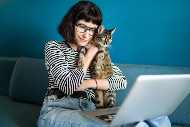 Jovem mulher trabalhando com laptop e com um gato sentado na moderna sala de estar em casa.