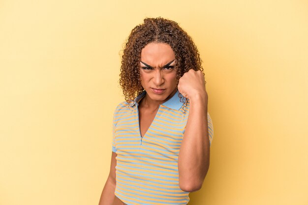 Foto jovem mulher transexual latina isolada em fundo amarelo, mostrando o punho para a câmera, expressão facial agressiva.