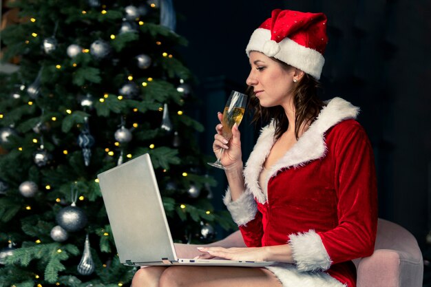 Jovem mulher triste com chapéu de papai noel com taça de champanhe, se comunicando através de um link de vídeo de laptop. conceito de natal, ano novo, comunicação online.