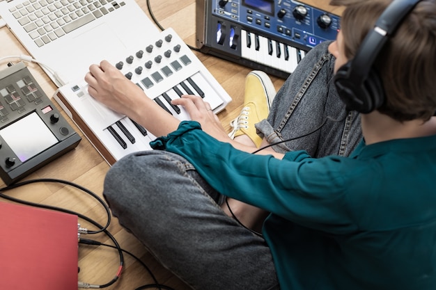 Jovem mulher usando fones de ouvido de estúdio, produzindo música eletrônica moderna. Músico feminino em home studio com instrumentos, laptop e processadores de efeitos.