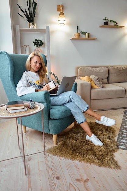 Jovem mulher usando laptop e escreve em um notebook sentado na poltrona trabalho remoto em casa