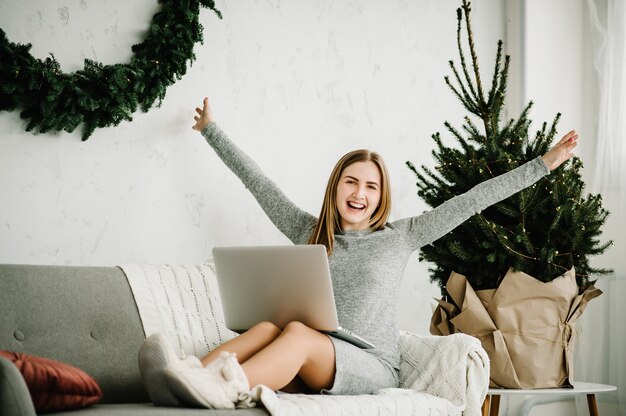Jovem mulher usando laptop e trabalhando em casa, perto de um pinheiro decorado com interior de Natal. Vendas de Natal. Menina digitando no laptop em casa. Conceito de Natal. Planejando feriados. Pesquisa na internet.