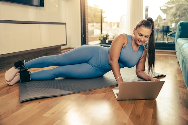 Jovem mulher usando laptop enquanto faz exercícios de fitness em casa pela manhã.