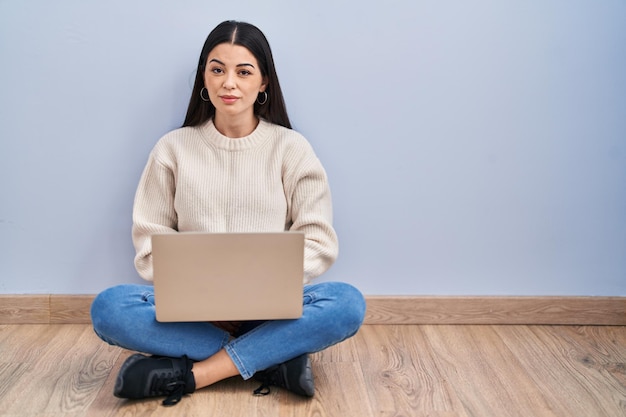 Jovem mulher usando laptop sentado no chão em casa relaxado com expressão séria no rosto simples e natural olhando para a câmera