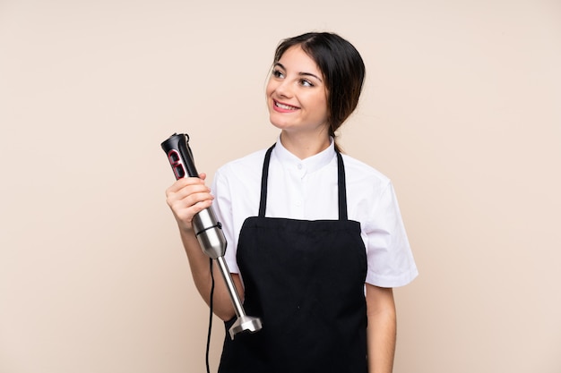 Jovem mulher usando liquidificador mão sobre parede, olhando para cima enquanto sorrindo