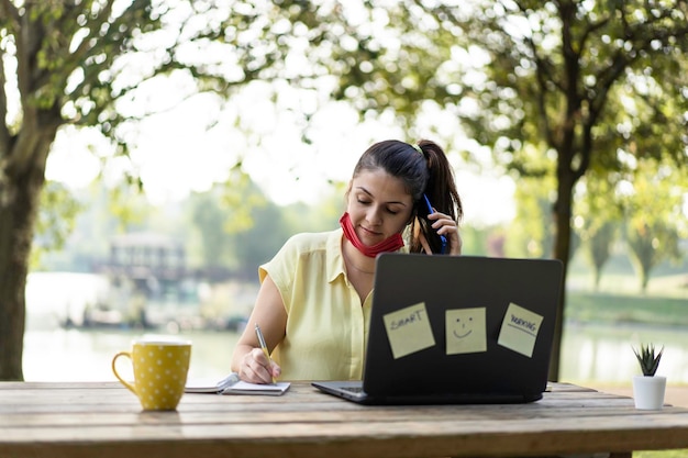 Jovem mulher usando máscara facial aberta chamando com smartphone enquanto trabalhava com laptop ao ar livre sorrindo freelancer feminina falando no celular e usando o pc no parque durante o final do surto de covid19