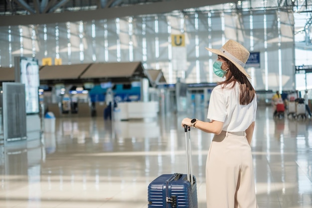 Jovem mulher usando máscara facial com bagagem andando na proteção do aeroporto infecção por doença de coronavírus viajante de mulher asiática com chapéu hora de viajar após o conceito de dose de reforço de vacina