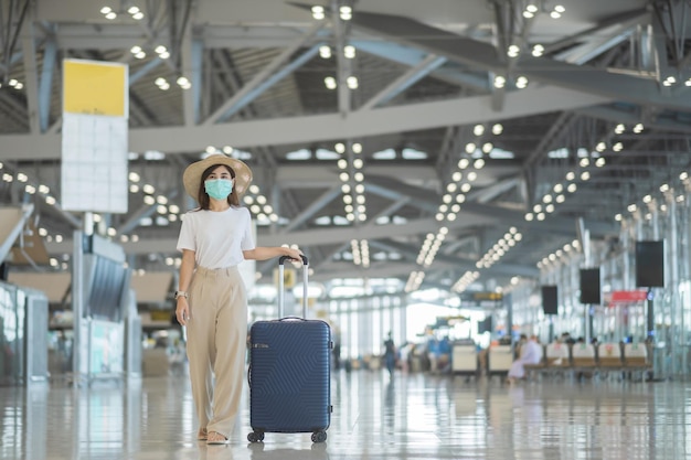 Jovem mulher usando máscara facial com bagagem andando na proteção do aeroporto Infecção por doença de coronavírus Viajante de mulher asiática com chapéu Hora de viajar após o conceito de dose de reforço de vacina