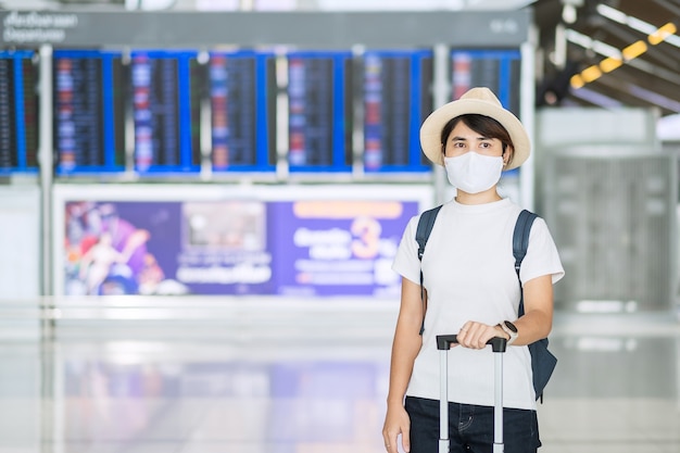 Jovem mulher usando máscara facial com bagagem, verificando o tempo de voo no aeroporto