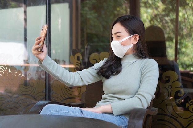 Jovem mulher usando máscara médica fazendo selfie com smartphone em um café, novo estilo de vida normal