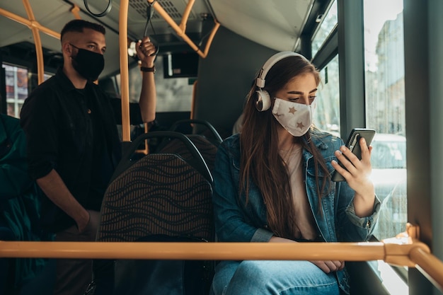 Jovem mulher usando máscara protetora e usando smartphone enquanto andava de ônibus