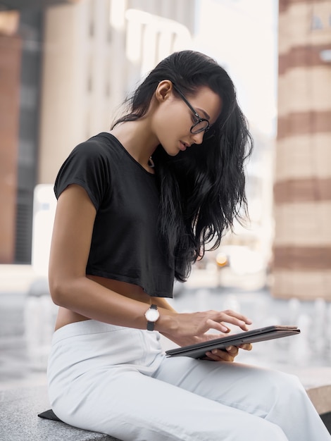 Jovem mulher usando o tablet pc na rua da cidade. Mulher de negócios da moda de óculos, trabalhando ao ar livre