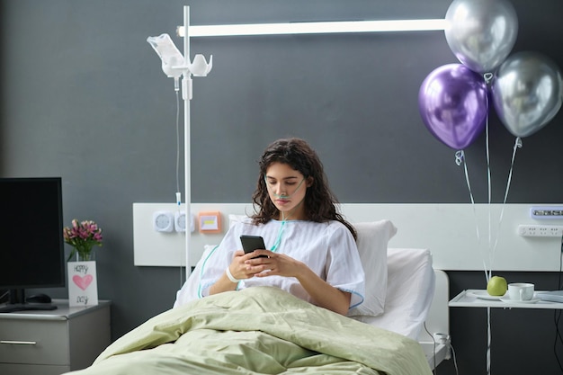 Jovem mulher usando seu smartphone enquanto estava deitado na cama na enfermaria do hospital decorada com balões