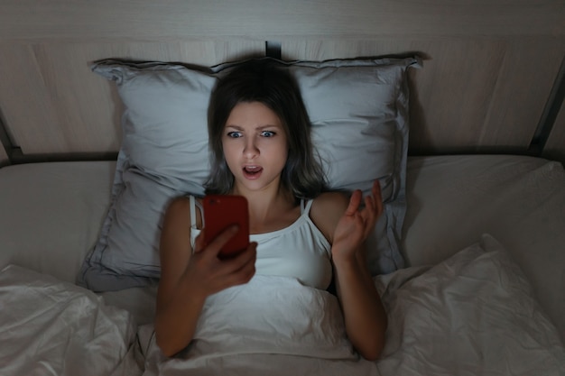 Jovem mulher usando seu telefone para navegar na Internet na cama à noite.