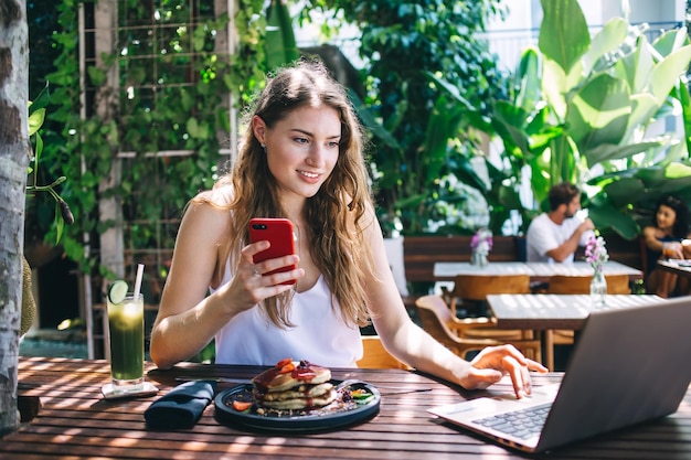 Jovem mulher usando smartphone e laptop no terraço do café