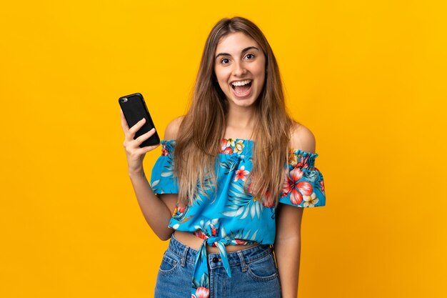 Jovem mulher usando telefone celular isolado parede amarela com surpresa e expressão facial chocado