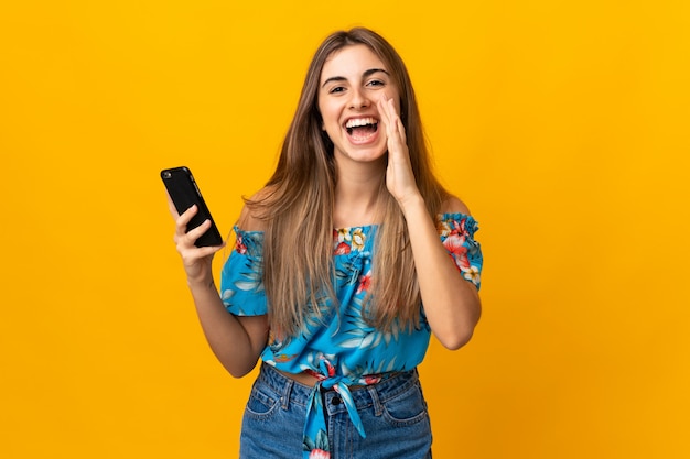 Jovem mulher usando telefone celular sobre parede amarela gritando com a boca aberta