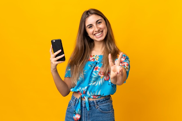 Jovem mulher usando telefone celular sobre parede amarela sorrindo e mostrando sinal de vitória