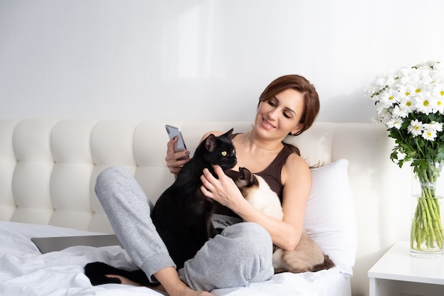 Jovem mulher usando telefone com seus dois gatos sentados juntos no quarto com flores.