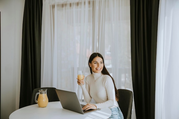 Jovem mulher usando um laptop e bebendo suco de laranja saudável no quarto