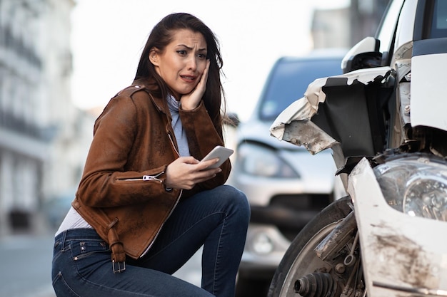 Jovem mulher usando um telefone celular enquanto se sente chocada com danos no carro após um acidente