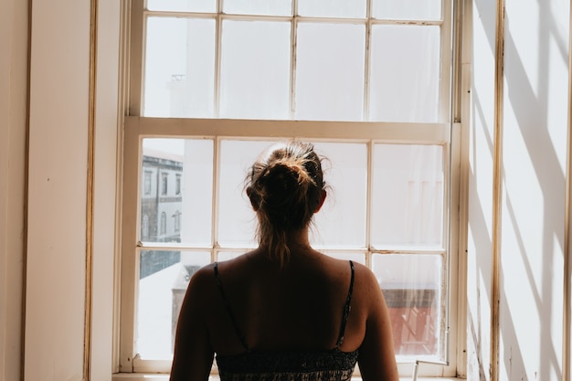 Jovem mulher usando um vestido olhando por uma janela, dando as costas para a câmera, conceitos de estresse e ansiedade, imagem escura, tristeza
