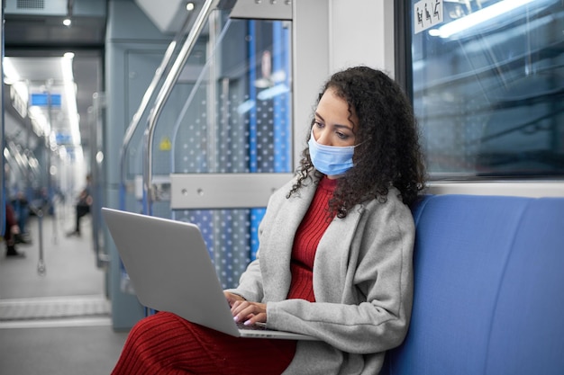 Jovem mulher usando uma máscara protetora usando um laptop durante uma viagem de metrô