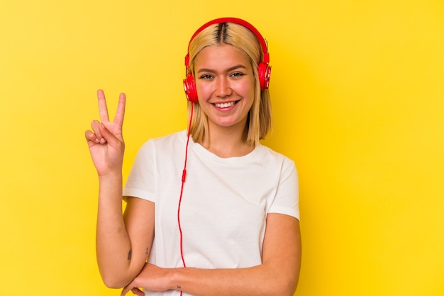 Jovem mulher venezuelana ouvindo música isolada na parede amarela, mostrando o número dois com os dedos.