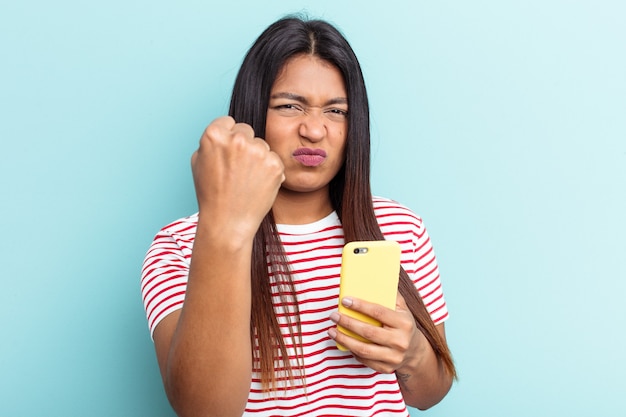 Jovem mulher venezuelana segurando celular isolado sobre fundo azul, mostrando o punho para a câmera, expressão facial agressiva.