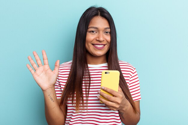 Jovem mulher venezuelana segurando celular isolado sobre fundo azul, sorrindo alegre mostrando o número cinco com os dedos.