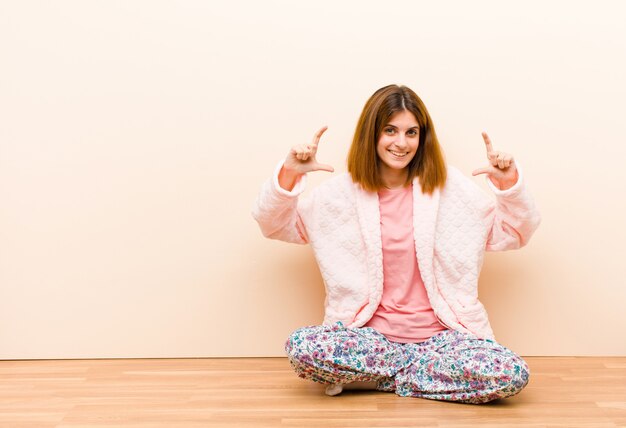 Jovem mulher vestindo pijama sentado em casa enquadrando ou descrevendo o próprio sorriso com as duas mãos, olhando o conceito de bem-estar positivo e feliz