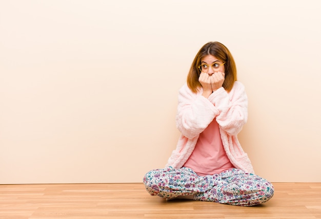 Jovem mulher vestindo pijama, sentado em casa olhando preocupado, ansioso, estressado e com medo, roer unhas e olhando para o espaço lateral da cópia