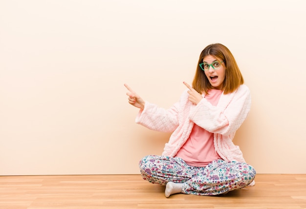 Jovem mulher vestindo pijama, sentado em casa, sentindo-se alegre e surpreso, sorrindo com uma expressão chocada e apontando para o lado