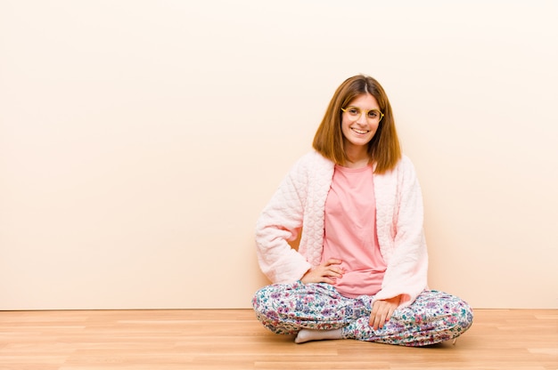 Jovem mulher vestindo pijama, sentado em casa, sorrindo alegremente com uma mão no quadril e atitude confiante, positiva, orgulhosa e amigável