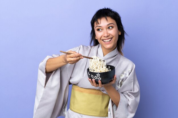 Jovem mulher vestindo quimono azul isolado, segurando uma tigela de macarrão com pauzinhos e olhando para cima