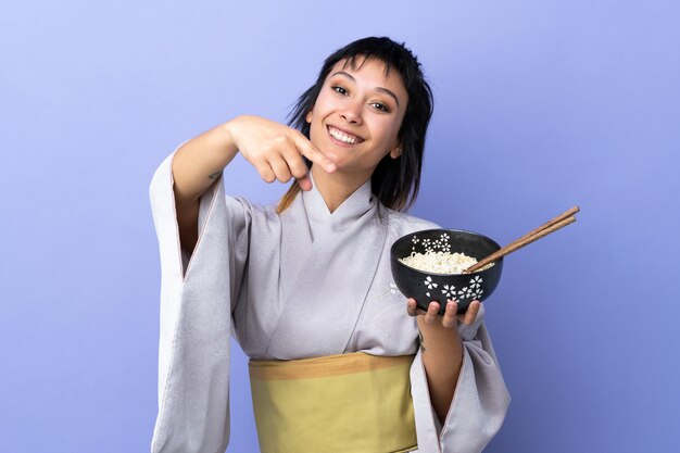 Jovem mulher vestindo quimono sobre dedo isolado azul pontos para você com uma expressão confiante, mantendo uma tigela de macarrão com pauzinhos