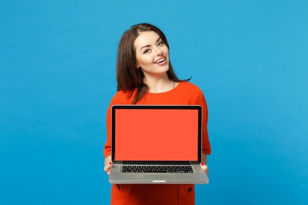 Jovem mulher vestindo vestido laranja vermelho segurar nas mãos usando computador portátil pc com tela vazia em branco isolada em fundo azul, retrato de estúdio. conceito de moda de estilo de vida de pessoas. mock up espaço de cópia.