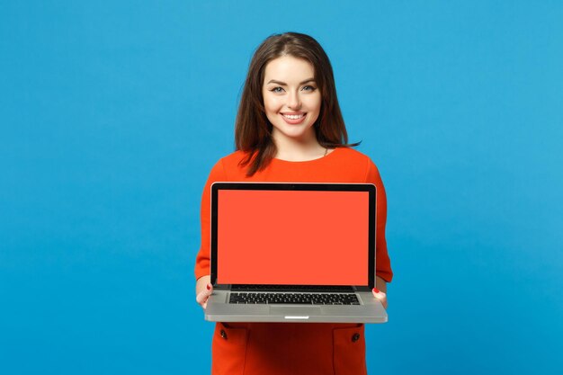 Jovem mulher vestindo vestido laranja vermelho segurar nas mãos usando computador portátil pc com tela vazia em branco isolada em fundo azul, retrato de estúdio. Conceito de moda de estilo de vida de pessoas. Mock up espaço de cópia.