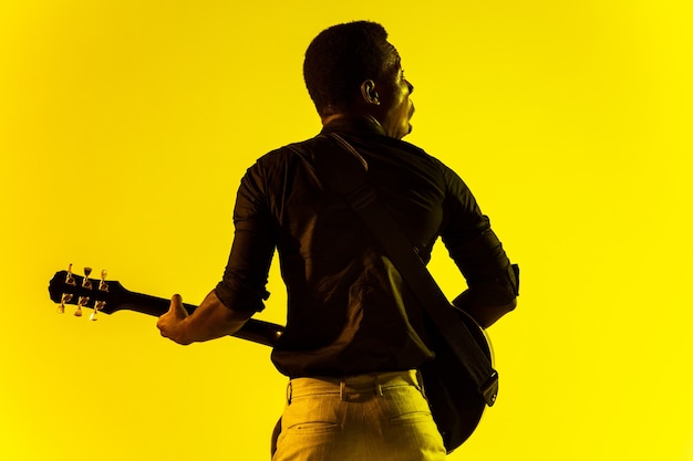 Jovem músico afro-americano tocando violão como uma estrela do rock na parede amarela em luz de néon.