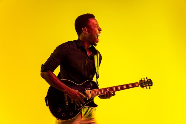 Jovem músico afro-americano tocando violão como uma estrela do rock sobre fundo amarelo em luz de néon. Conceito de música, hobby, festival, ao ar livre. Cara alegre e atraente improvisando, cantando.