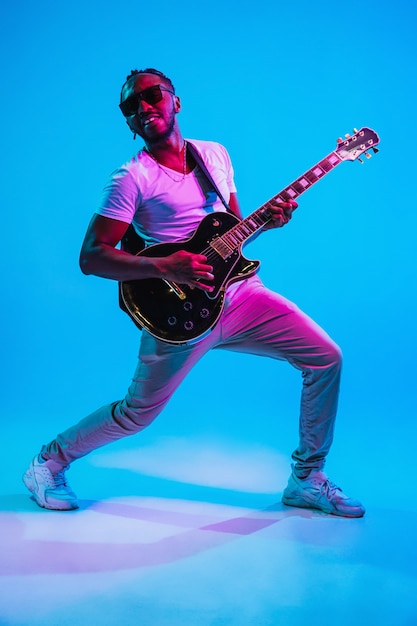 Jovem músico afro-americano tocando violão como uma estrela do rock sobre fundo azul do estúdio em luz de néon. conceito de música, hobby. cara alegre improvisando. retrato retro colorido.