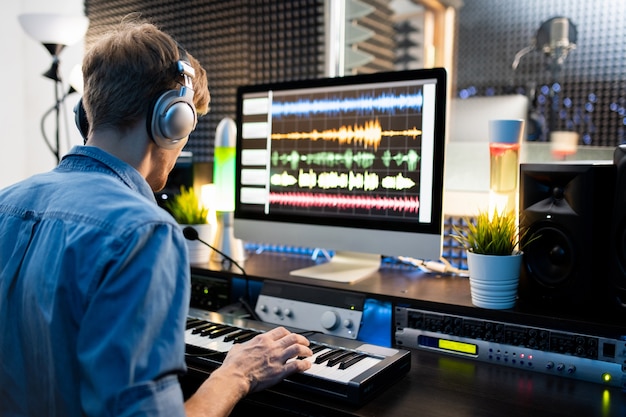 Jovem músico com fones de ouvido pressionando as teclas do teclado de piano enquanto está sentado ao lado do monitor do computador e trabalhando com sons