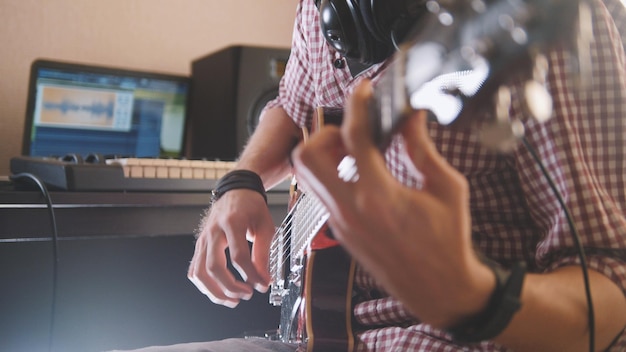 Jovem músico compõe e grava música tocando guitarra elétrica, usando computador e teclado, foco nas mãos