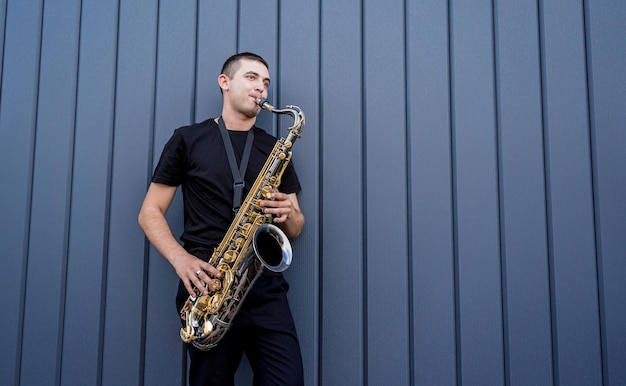 Jovem músico de rua tocando saxofone perto da grande parede azul