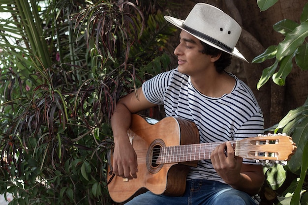 Jovem músico hispânico gosta de tocar violão ao ar livre entre as plantas no jardim, ele gosta muito de seu hobby e a expressão em seu rosto confirma isso