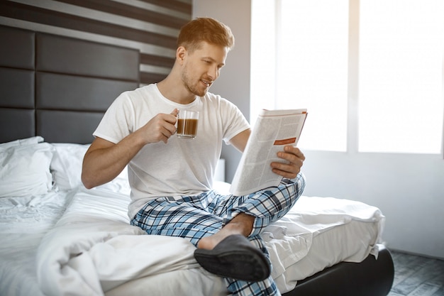 Foto jovem na cama de manhã. ele senta e lê o jornal guy, segurando uma xícara de café. agradável e alegre.