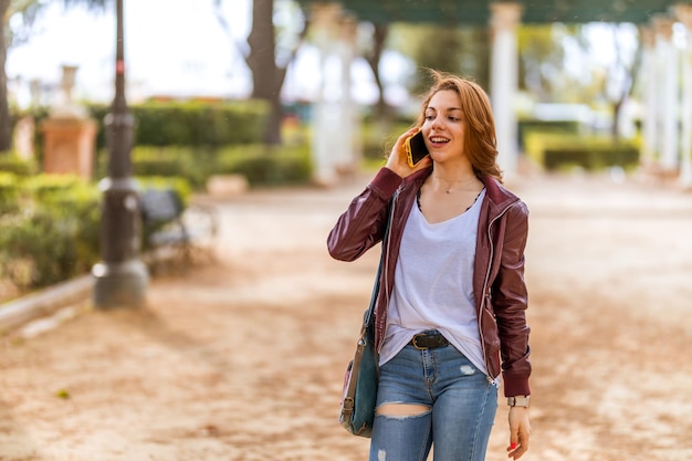 Jovem natural caminhando enquanto fala e sorrindo com seu smartphone no parque