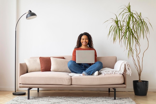 Jovem negra alegre trabalhando em seu laptop enquanto estava sentada no sofá