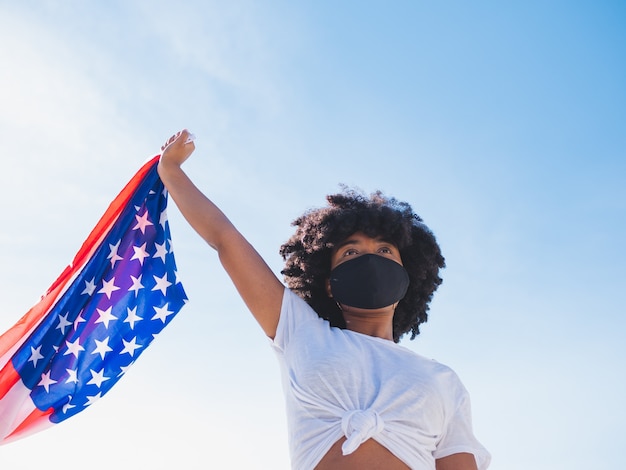 Jovem negra ao ar livre, usando uma máscara protetora contra o coronavírus e segurando uma bandeira dos eua